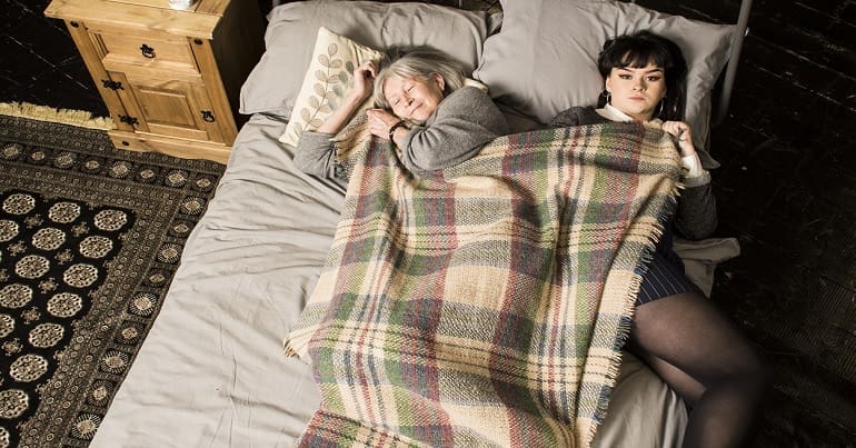 Promotional image from The Fall of an old woman sleeping in a bed beside a young woman who is awake and staring at the camera