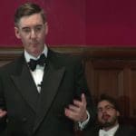 Jacob Rees-Mogg speaking while people talk behind him