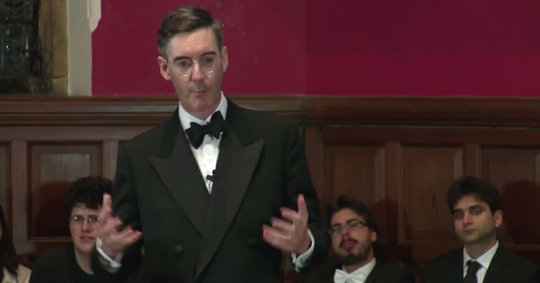 Jacob Rees-Mogg speaking while people talk behind him