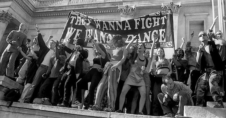 People dancing in Trafalgar Square with a banner that says 'They Wanna Fight, We Wanna Dance'