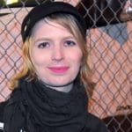 A picture of Chelsea Manning standing in front of a fence and raising her fist.