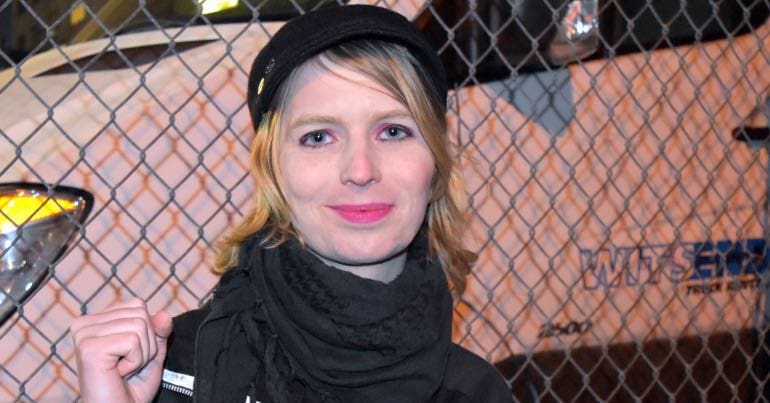 A picture of Chelsea Manning standing in front of a fence and raising her fist.