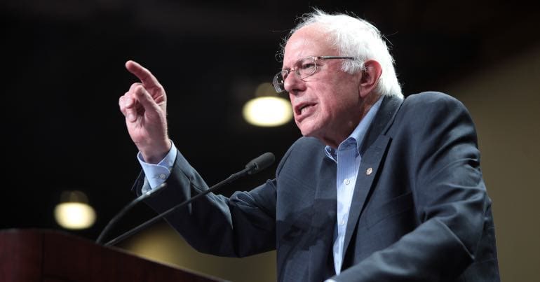 A picture of Bernie Sanders at a lectern pointing at someone or something.