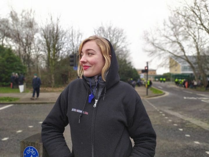 Isabel a geography student from Germany on looking a Yellow Vest member greting traffic in the middle of the road with a pro Assange poster encampment a short with Belmarsh behind her