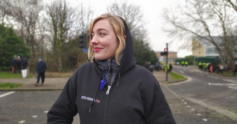 Isabel a geography student from Germany on looking a Yellow Vest member greting traffic in the middle of the road with a pro Assange poster encampment a short with Belmarsh behind her