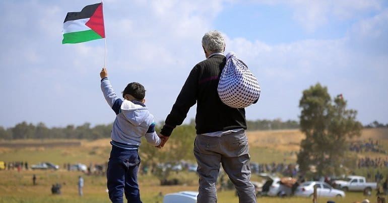 A Palestinian child holds up the country's flag while holding the hand of an older man with a sack drapped over his shoulder. They are stood on a raised dirt hill overlooking a landscape where others are gathered.