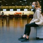 Woman at airport wearing face mask, sitting on her luggage
