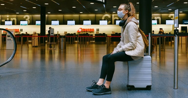 Woman at airport wearing face mask, sitting on her luggage