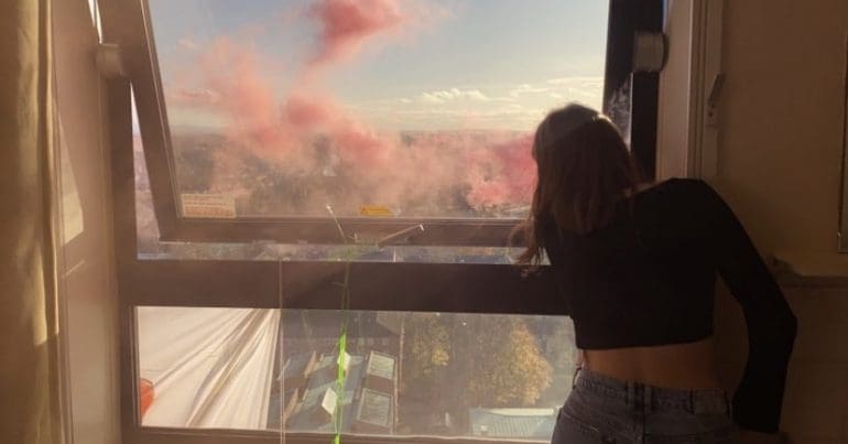 A student protestor looking out at Manchester from an occupied high rise window as coloured flare smoke fills the air