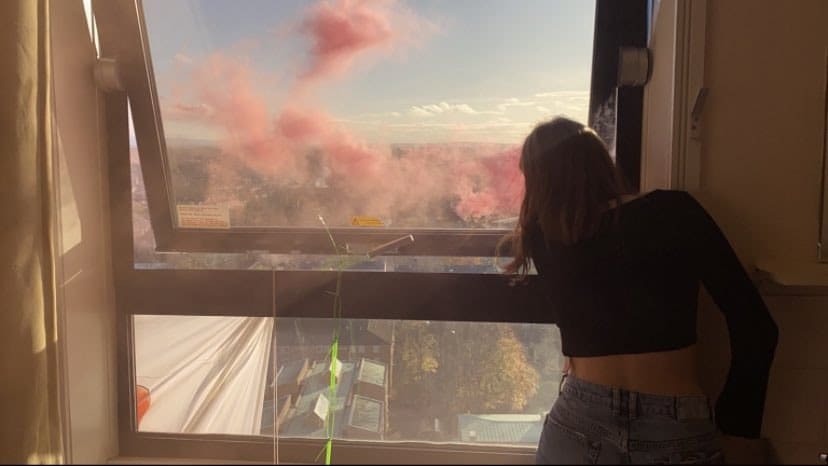A student protestor looking out at Manchester from an occupied high rise window as coloured flare smoke fills the air