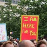 A crowd of people marching in front of trees, with a large old-fashioned building in the background. You can only see the tops of people's heads, and two placards. One placard says in multicoloured letters 'Love is tender and knows no gender' with 'out' and 'proud' in smaller letters. The other placard says 'I am here for you'.
