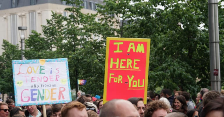 A crowd of people marching in front of trees, with a large old-fashioned building in the background. You can only see the tops of people's heads, and two placards. One placard says in multicoloured letters 'Love is tender and knows no gender' with 'out' and 'proud' in smaller letters. The other placard says 'I am here for you'.