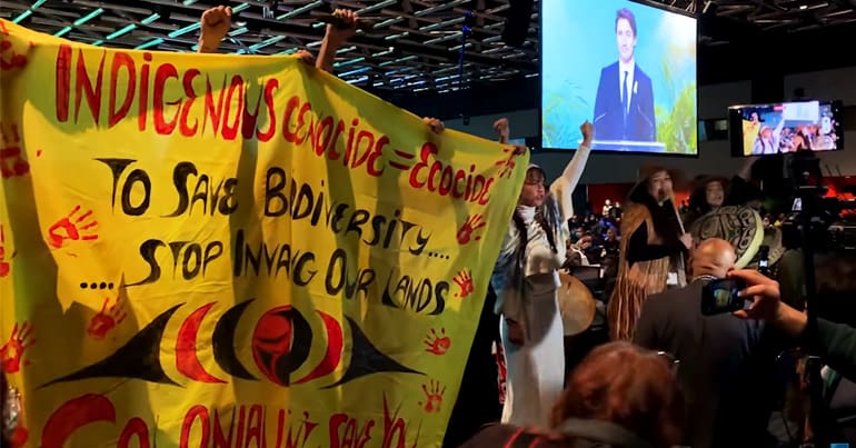 Indigenous youth hold up a banner reading "indigenous genocide = ecocide", disrupting a speech by Justin Trudeau at the opening of COP15