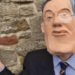 Someone wearing a Jacob Rees Mogg mask pointing at a satirical blue plaque