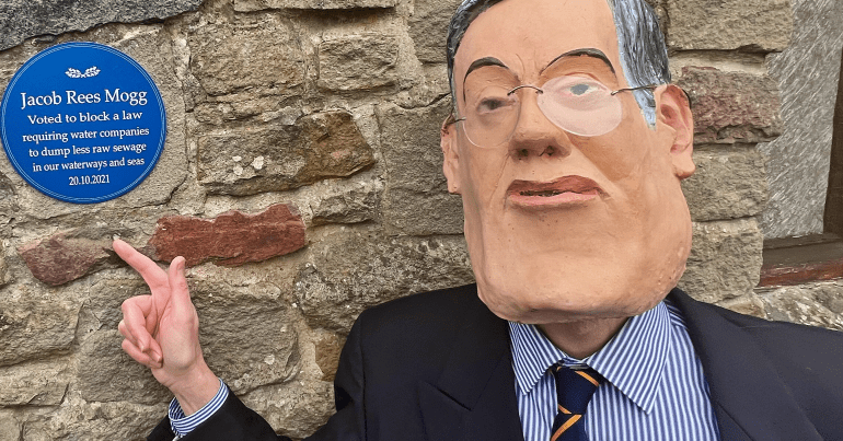 Someone wearing a Jacob Rees Mogg mask pointing at a satirical blue plaque