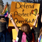 an individual holds a banner reading "defend & protect queer kids"