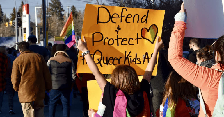 an individual holds a banner reading "defend & protect queer kids"