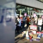 An NHS sign is blurred in the foreground with a picket line in focus in the background