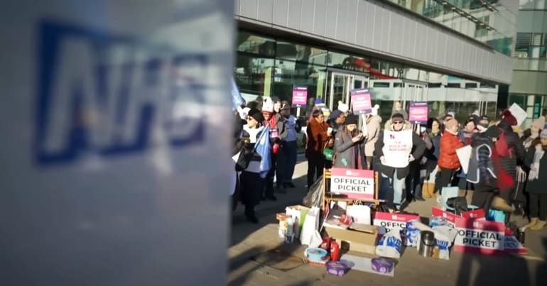 An NHS sign is blurred in the foreground with a picket line in focus in the background