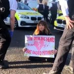 An Animal Rebellion protester sits on the road holding a sign saying "stop breaking hearts - be part of a plast-based future"