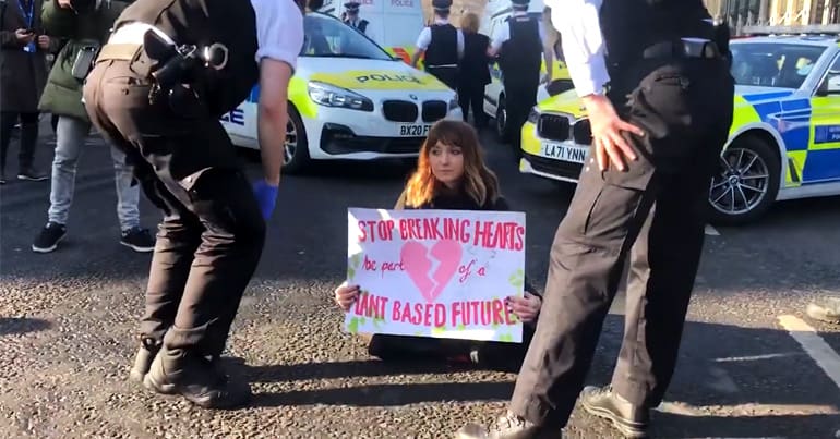 An Animal Rebellion protester sits on the road holding a sign saying "stop breaking hearts - be part of a plast-based future"