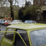 Rowan Atkinson as Mr Bean stood by his yellow car. Guardian EVs Electric Vehicles