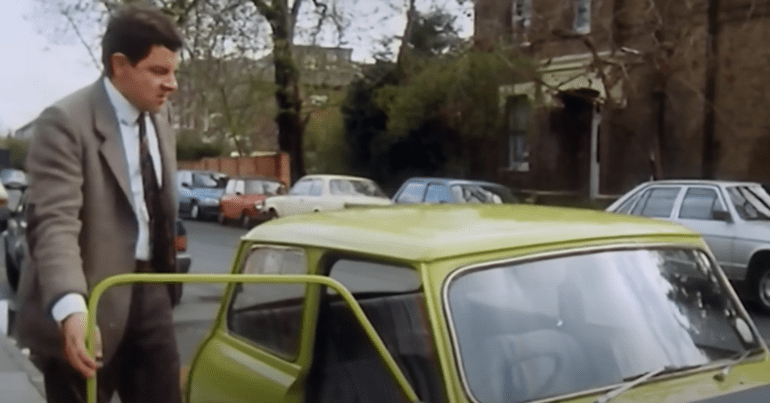 Rowan Atkinson as Mr Bean stood by his yellow car. Guardian EVs Electric Vehicles