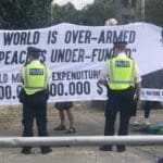 Image is of a banner at DSEI 2019 saying "the world is over-armed and peace is under-funded" with cops standing in front of it.