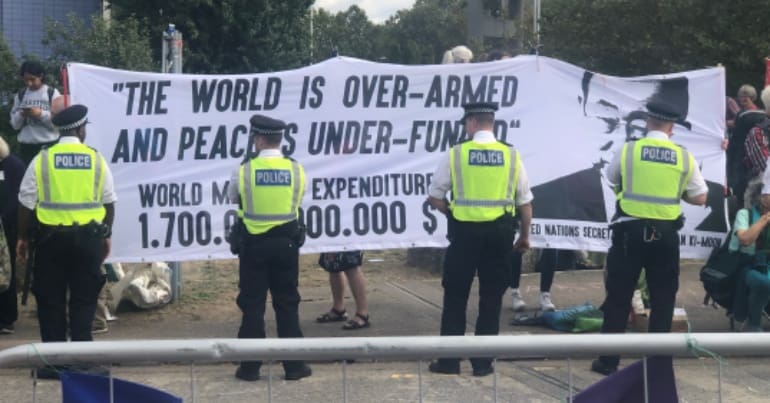 Image is of a banner at DSEI 2019 saying "the world is over-armed and peace is under-funded" with cops standing in front of it.