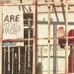Palestinian woman stands behind wire fence, next to a sign that reads "Arabs are prohibited. This is apartheid."