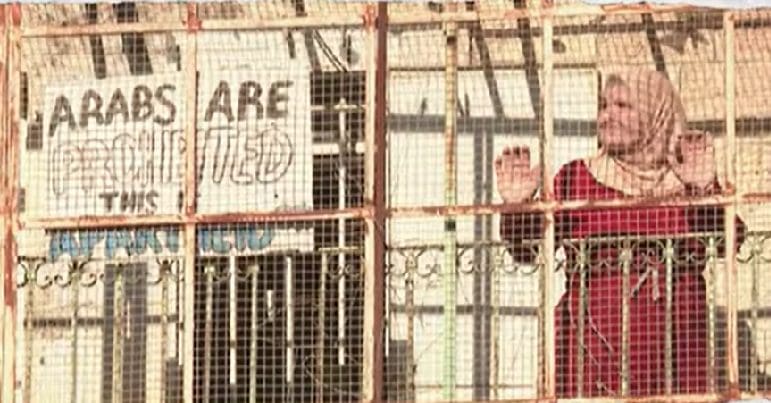 Palestinian woman stands behind wire fence, next to a sign that reads "Arabs are prohibited. This is apartheid."