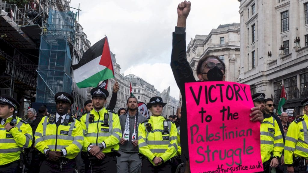 Black woman with a raised fist stands in front of a line of police holding a sign that says 'Victory to the Palestinian struggle'
