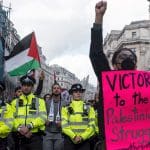 Black woman with a raised fist stands in front of a line of police holding a sign that says 'Victory to the Palestinian struggle'