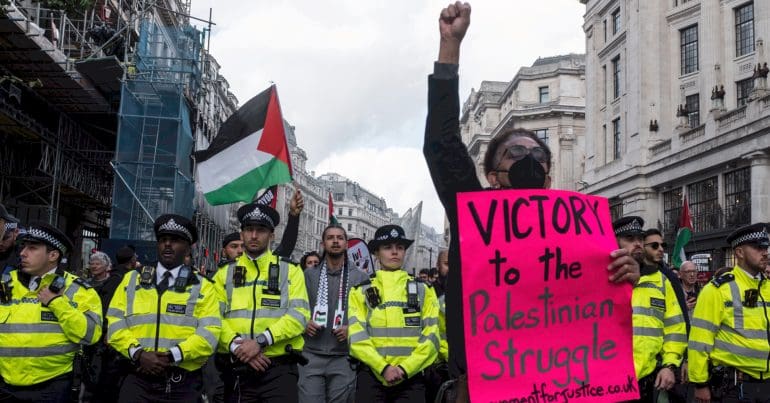 Black woman with a raised fist stands in front of a line of police holding a sign that says 'Victory to the Palestinian struggle'