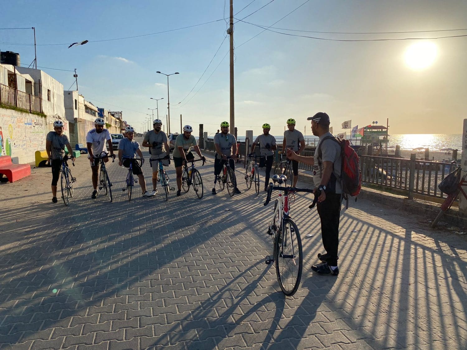 The Gaza Sunbirds on their bikes, stationary and lined up. It is nearly sunset. You can see the Mediterranean sea in the background. Their team leader is standing talking to them. 