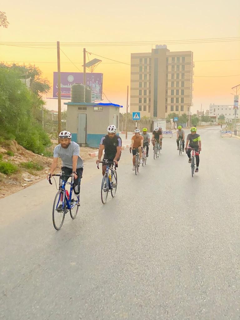 The Gaza Sunbirds cycling on a road. There is a tower block in the background and it is morning as the sun is coming up in the distance. 