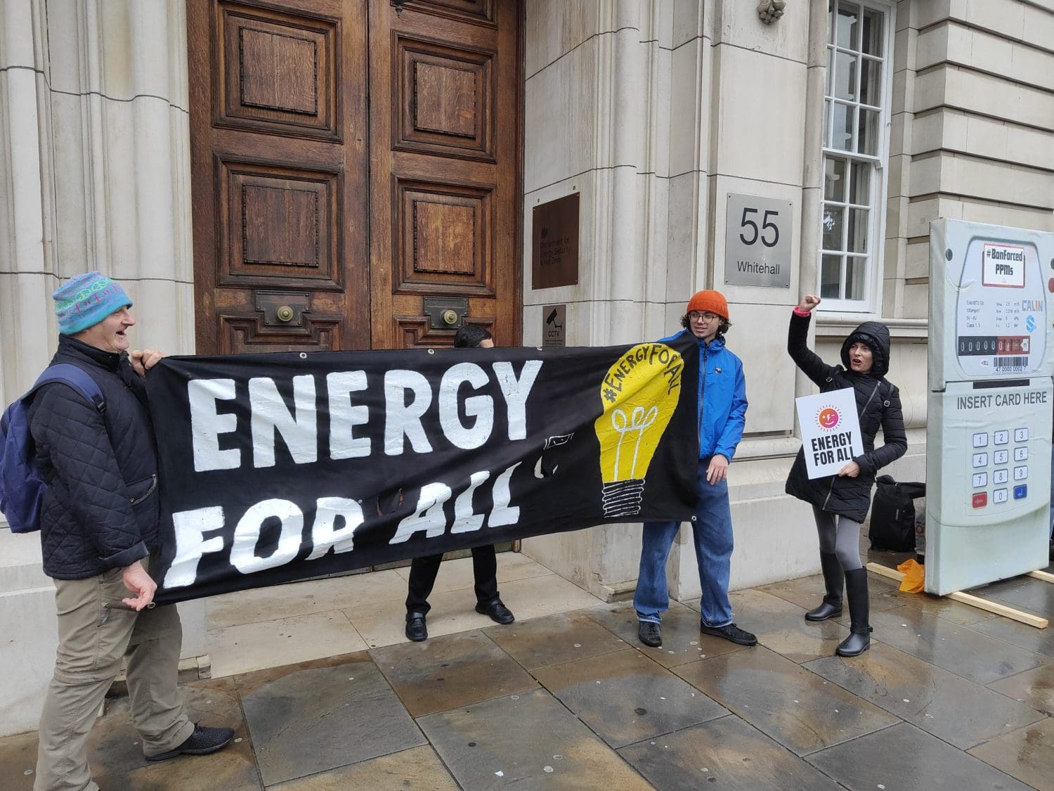 Protesters holding a banner that says "energy for all" and a placard that says the same outside a big wooden door