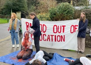 A Bristol University Arms Industry Blood out of Education protest in 2024. A student kneels on the ground covered in red liquid and another lies motionless. ‘Blood out of education’ is written in red on a banner behind them.