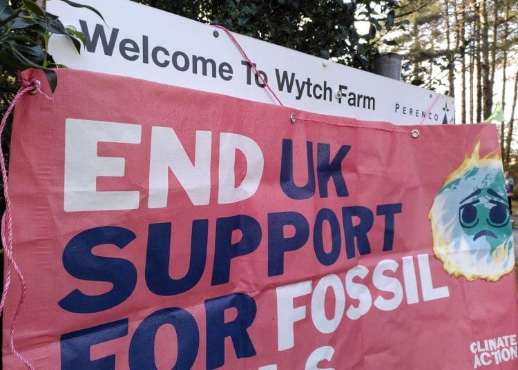 Entrance sign to Perenco's onshore oilfield which reads "Welcome to Wytch Farm", while a sign reading: "End UK support for fossil fuels" with a burning earth image is draped over the rest.