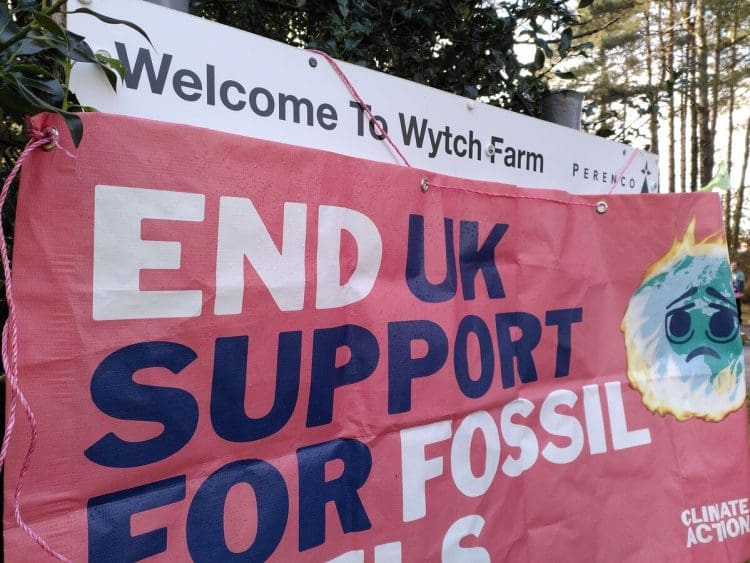 Entrance sign to Perenco's onshore oilfield which reads "Welcome to Wytch Farm", while a sign reading: "End UK support for fossil fuels" with a burning earth image is draped over the rest.