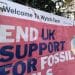 Entrance sign to Perenco's onshore oilfield which reads "Welcome to Wytch Farm", while a sign reading: "End UK support for fossil fuels" with a burning earth image is draped over the rest.