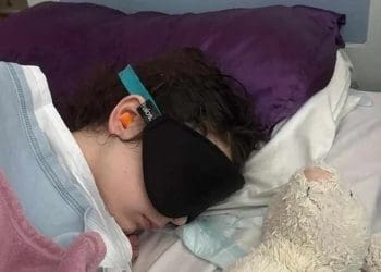 Millie in hospital wearing a face and ear coverings wrapped in a blanket severe ME