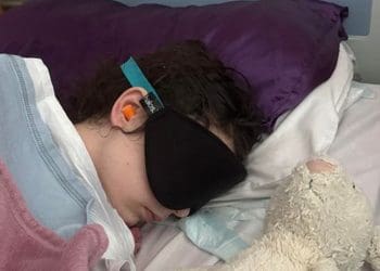 Millie McAinsh lying in bed wearing ear protectors and an eye mask with a teddy lying next to her ME/CFS