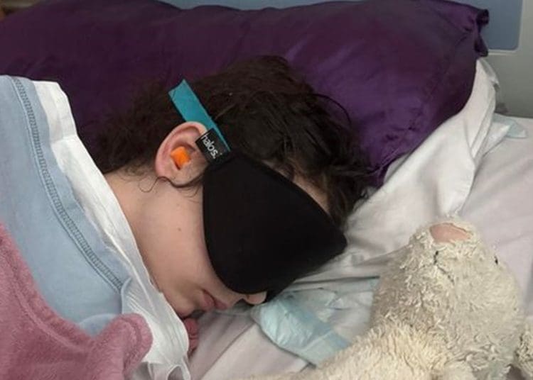 Millie McAinsh lying in bed wearing ear protectors and an eye mask with a teddy lying next to her ME/CFS