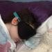 Millie McAinsh lying in bed wearing ear protectors and an eye mask with a teddy lying next to her ME/CFS