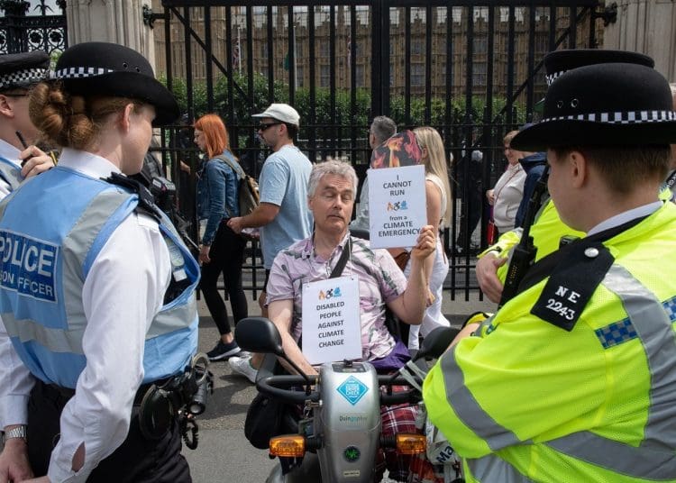 Disabled man Neil Goodwin protesting in his mobility scooter at the entrance to parliament. He is surrounded by police officers and holding signs that read "disabled people against climate change" and ""I cannot run from a climate emergency"