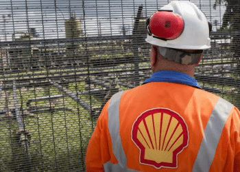 Shell worker with tabard emblazoned with Shell's logo looks out through a fence at oil and gas infrastructure Nigeria