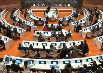 UNCRPD forum room at Geneva there are circles of long tables with people sitting at screens at the top of the room is a long table against a marble stone backdrop