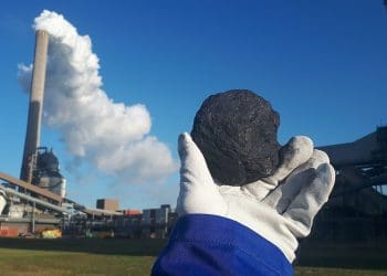 Image shows piece of coal being held in front of coal power plant