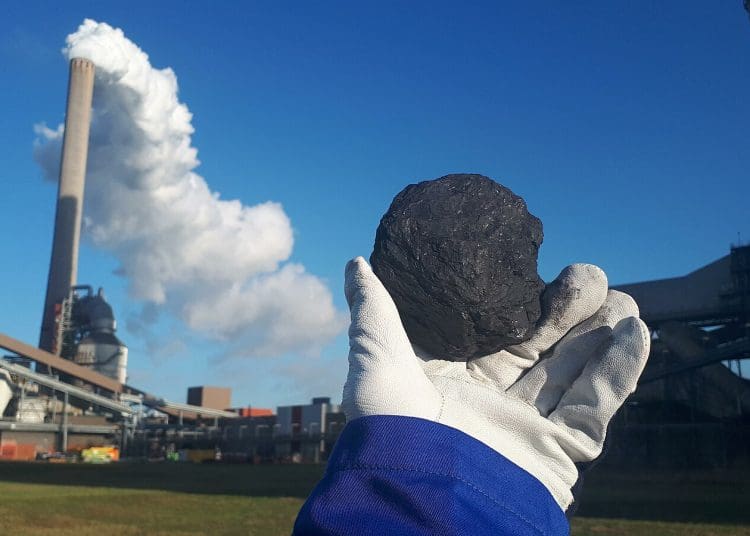Image shows piece of coal being held in front of coal power plant